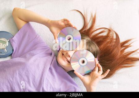 Happy teenage girl in lilac t-shirt lies on bed in pink headphones holds cd disks in her hands Stock Photo