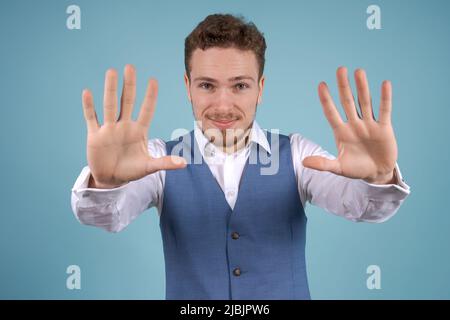 Portrait young business man with crossed arms, looking at camera, on blue background. Bearded guy in white shirt and blue vest posing in studio Stock Photo