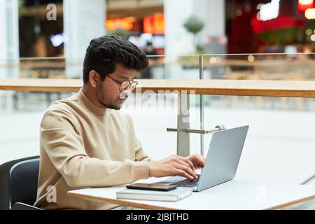Side view portrait of young man of Indian ethnicity using laptop in shopping mall cafe, copy space Stock Photo
