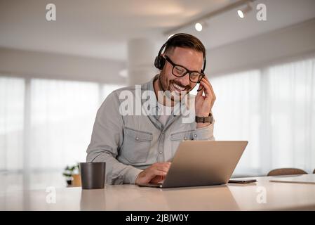 Young smiling businessman video conferencing on laptop. Happy professional discussing through headset at desk. He is working in corporate office. Stock Photo