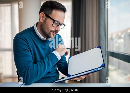 Focused young businessman analyzing documents. Serious professional is wearing smart casuals. He is working at desk in office. Stock Photo