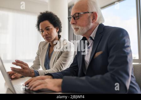 Serious male and female business partners working on laptop. Professionals are planning strategy during meeting. They are sitting at desk in office. Stock Photo