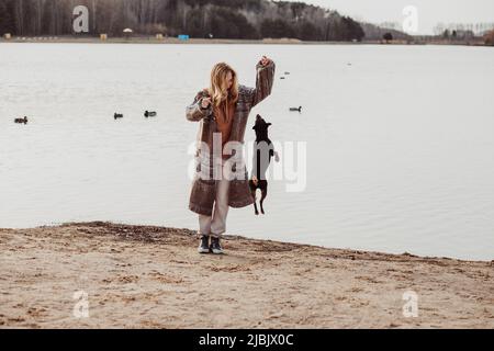 Happy enjoying blonde woman in warm clothes playing and having fun with cute puppy near river bank on cool sunny day, ducks swimming in water Stock Photo