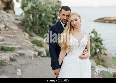 Portrait of young blonde bride in white dress and brunet groom in suit embracing and holding hands on rocky sea coast in Italy, seascape background Stock Photo