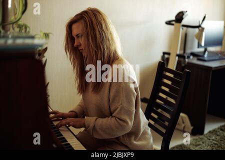 Beautiful blonde woman in home clothes sitting at piano, pressing keys and playing melody side view. Young lady practice playing musical instrument at Stock Photo