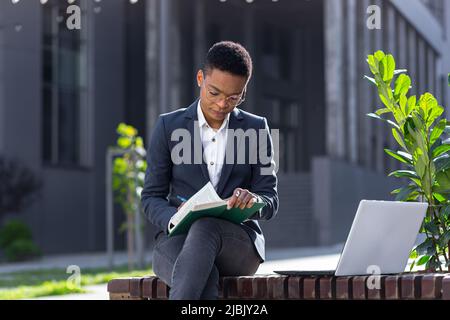 Young beautiful female student studying, remotely in the park, sitting on a bench, online and remotely, using laptop, near college and campus Stock Photo