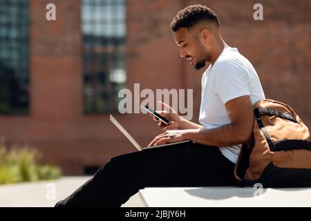 Young african american guy with laptop sitting outdoors and using smartphone, surfing internet or texting, side view Stock Photo