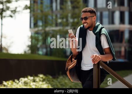 On my way to work. Happy stylish african american man using his smartphone and walking in park area, free space Stock Photo