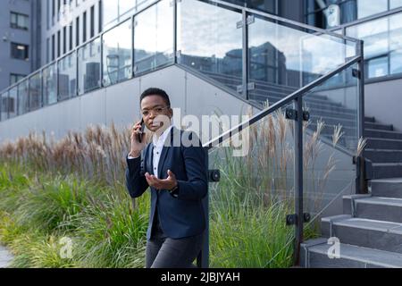 Angry and serious businesswoman talking on the phone outside the office, African American woman dressed in business clothes, communicating with collea Stock Photo