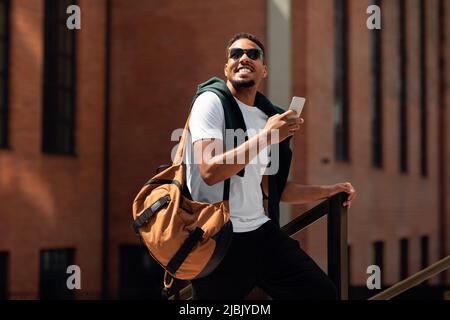Happy trendy black guy in sunglasses using cellphone while walking down the street in urban city, copy space Stock Photo