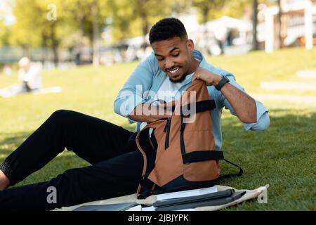 Happy black man searching something in his backpack, sitting on grass in park outdoors Stock Photo