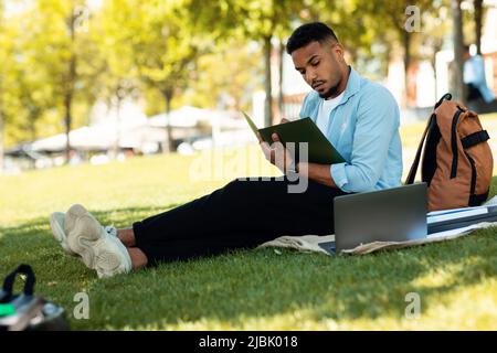E-learning concept. African american male student preparing for lecture, using laptop and taking notes, sitting in park Stock Photo
