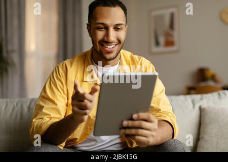 Happy young black man with tablet sitting on couch, working or studying online, having video chat at home Stock Photo