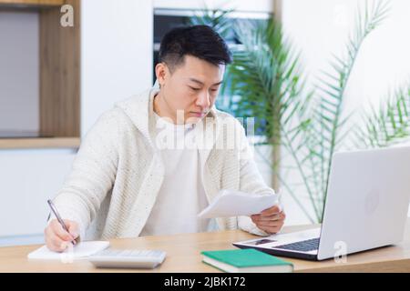 Young Asian man doing paperwork at home sitting at a desk, working on a laptop, focused on paying bills Stock Photo