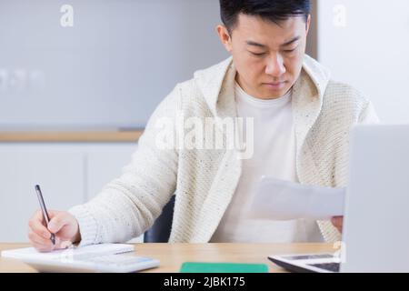 Young Asian man doing paperwork at home sitting at a desk, working on a laptop, focused on paying bills Stock Photo
