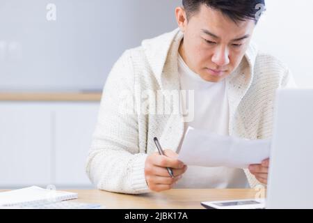 Young Asian man doing paperwork at home sitting at a desk, working on a laptop, focused on paying bills Stock Photo