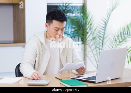 Young Asian man doing paperwork at home sitting at a desk, working on a laptop, focused on paying bills Stock Photo