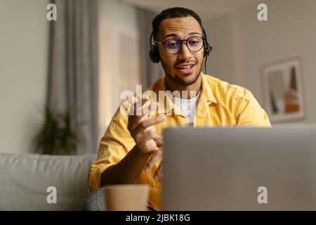Young African American man in headset having web call, using laptop, sitting on sofa, talking at webcam in home office Stock Photo