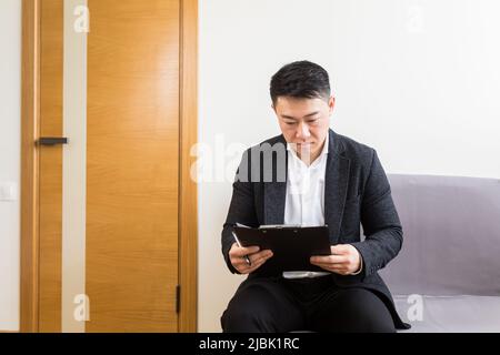 Young successful Asian man, waiting for a job interview, in the waiting room of the office center, sitting on a chair near the reception of the office Stock Photo