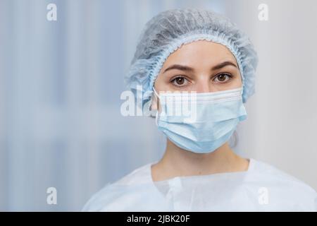 Close-up portrait of a young female doctor student in medical mask and surgical cap, in white coat, looking at camera Stock Photo