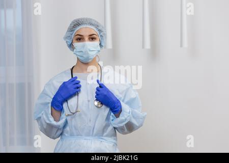 Portrait of young female doctor in mask, surgical gown and hat, standing holding stethoscope, looking at camera Stock Photo