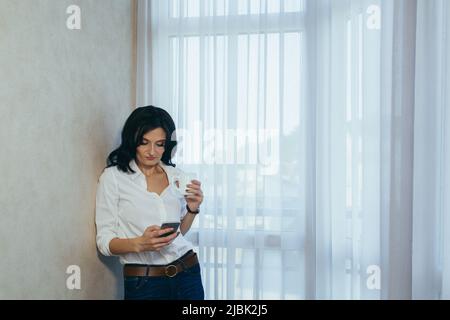 Beautiful young woman works studying at home, stands talking on the phone. Conducts business negotiations, talks with friends, drinks coffee Stock Photo