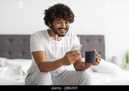 Cheerful eastern man drinking coffee and using smartphone Stock Photo