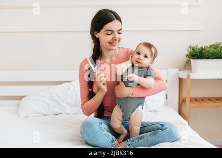 Happy mother combing hair of adorable baby girl, holding daughter on hands and smiling, little child looking at camera Stock Photo