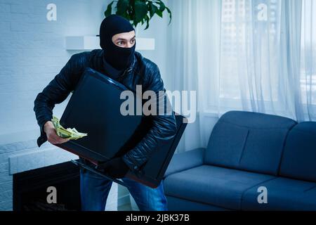 Robber man dressed in black hoodie stands with disguised face and holds a lot of money in his hands, stole a large amount, a thief man stole a TV Stock Photo