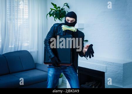 Robber man dressed in black hoodie stands with disguised face and holds a lot of money in his hands, stole a large amount, a thief man stole a TV Stock Photo
