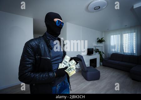 Robber man dressed in black hoodie stands with disguised face and holds a lot of money in his hands, stole a large amount, a thief man stole a TV Stock Photo
