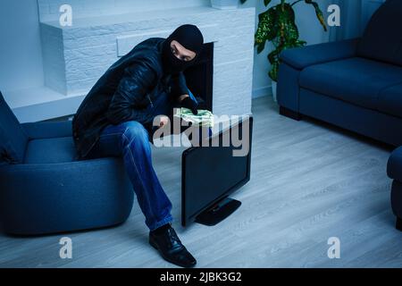 Robber man dressed in black hoodie stands with disguised face and holds a lot of money in his hands, stole a large amount, a thief man stole a TV Stock Photo