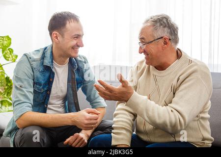 Smiling grown son sit on couch relax with senior dad talk sharing thoughts looking in eyes, happy millennial man rest on sofa speak with elderly Stock Photo
