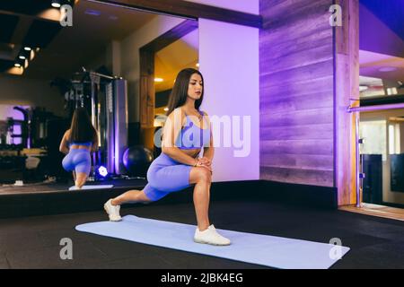 young beautiful sports woman in the gym doing exercises on a mat, in front of a mirror, arm swing, squats, fitness, yoga, in sportswear Stock Photo