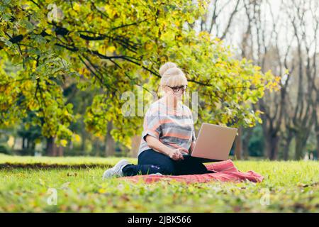Beautiful blonde woman, senior, sitting on the grass in the park uses a laptop, works, studies, talks on video link Stock Photo