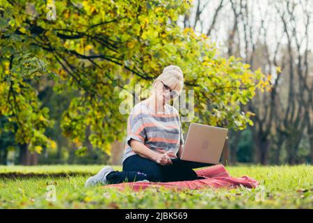 Beautiful blonde woman, senior, sitting on the grass in the park uses a laptop, works, studies, talks on video link Stock Photo