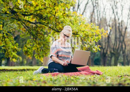 Beautiful blonde woman, senior, sitting on the grass in the park uses a laptop, works, studies, talks on video link Stock Photo