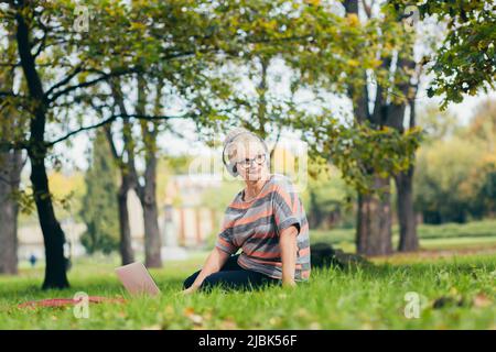 Beautiful blonde woman, senior, sitting on the grass in the park in headphones, uses a laptop, works, studies, listens to music, talks on the video pl Stock Photo