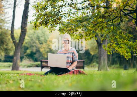 Beautiful blonde woman, senior, sitting on the grass in the park uses a laptop, works, studies, talks on video link Stock Photo