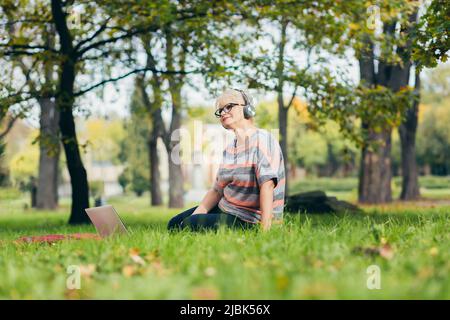 Beautiful blonde woman, senior, sitting on the grass in the park in headphones, uses a laptop, works, studies, listens to music, talks on the video pl Stock Photo