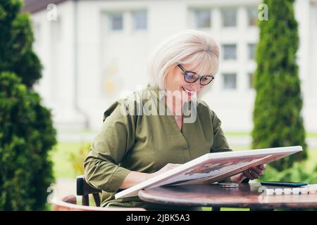 Senior beautiful blonde woman sitting at a table on the street, paints a picture, smiling, happy Stock Photo