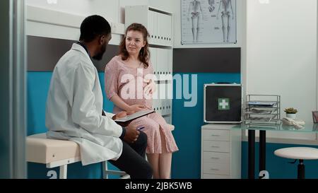 Male physician taking notes on checkup files at consultation with expectant patient, having conversation in medical office. Health specialist meeting with future mother at appointment. Stock Photo