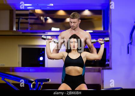 Young sports fitness trainer man trains young beautiful woman athlete in gym, shows exercises with dumbbells on biceps, legs Stock Photo