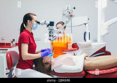 Woman dentist looks at the patient's teeth with a dental microscope and holds dental instruments near his mouth. The assistant helps the doctor with m Stock Photo