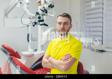 Portrait of a young dentist, assistant in a yellow medical uniform, sitting in a dental office, looking at the camera, arms crossed, smiling Stock Photo