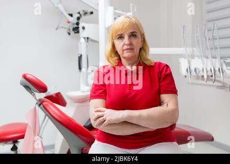 Portrait of a young beautiful female dentist, assistant in a red medical uniform, sitting in the dentist's office, looking at the camera, arms crossed Stock Photo