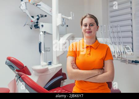 Portrait of a young beautiful female dentist, assistant in orange medical uniform, sitting in the dental office, looking at the camera, arms crossed, Stock Photo