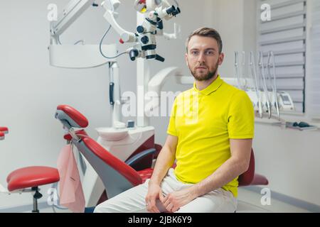 Portrait of a young dentist, assistant in a yellow medical uniform, sitting in a dental office, looking at the camera, arms crossed, smiling Stock Photo
