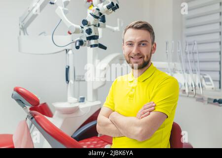 Portrait of a young dentist, assistant in a yellow medical uniform, sitting in a dental office, looking at the camera, arms crossed, smiling Stock Photo