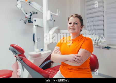 Portrait of a young beautiful female dentist, assistant in orange medical uniform, sitting in the dental office, looking at the camera, arms crossed, Stock Photo